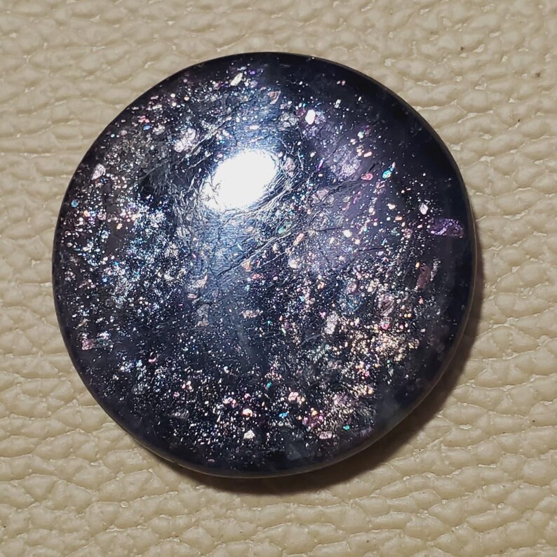 May include: A round, dark blue gemstone with a shimmering, speckled surface. The stone has a polished appearance with iridescent flecks of silver, purple, and green. The gemstone is set against a neutral background.