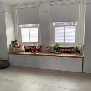 May include: A window seat with white trim and a wooden top, adorned with Christmas-themed pillows and plush toys. The pillows have festive text and imagery. The windows have blinds.