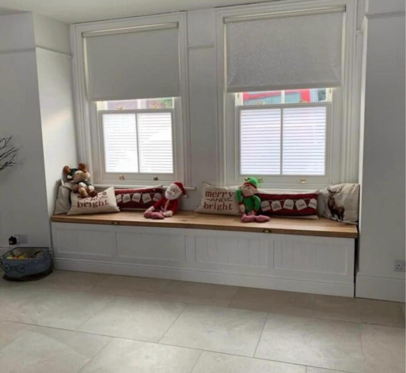 May include: A window seat with white trim and a wooden top, adorned with Christmas-themed pillows and plush toys. The pillows have festive text and imagery. The windows have blinds.