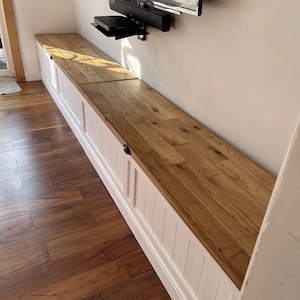 May include: A long, built-in storage bench with a light wood top and white paneled base. The bench runs along a wall, with a TV mounted above. The wood flooring complements the bench's design. The bench has a length of approximately 10 feet.