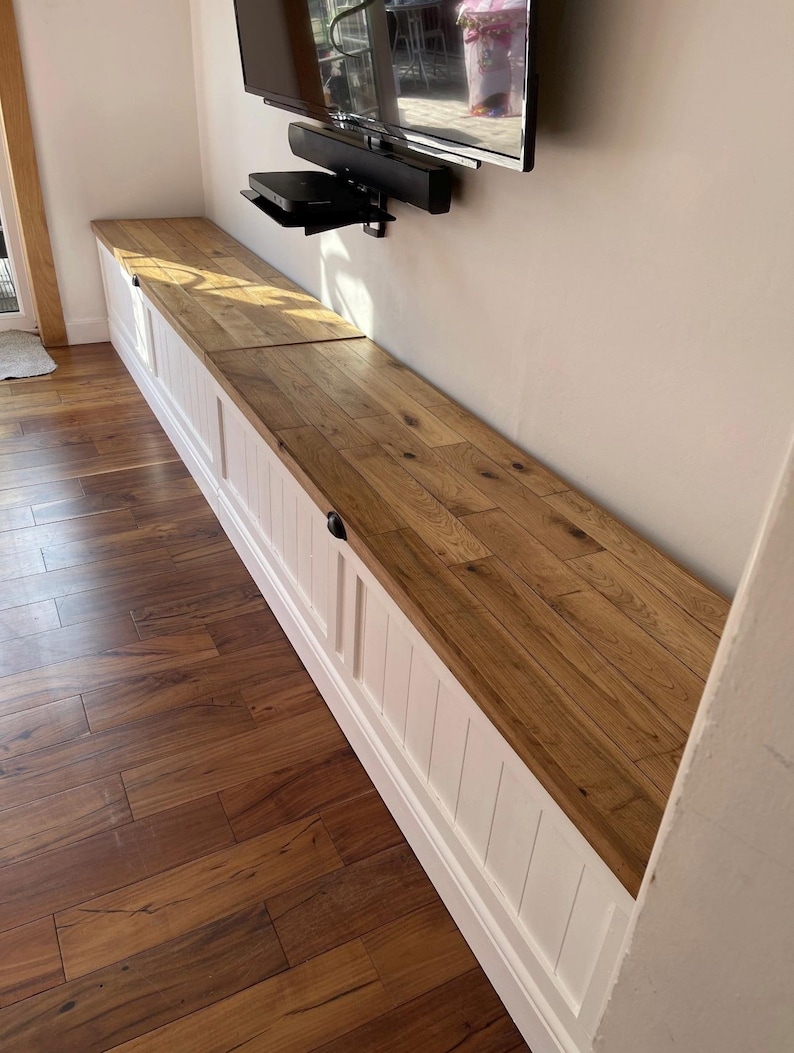 May include: A long, built-in storage bench with a light wood top and white paneled base. The bench runs along a wall, with a TV mounted above. The wood flooring complements the bench's design. The bench has a length of approximately 10 feet.