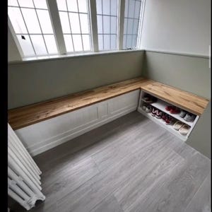 May include: A built-in wooden bench with a white base and a natural wood top. The bench is positioned beneath a large window and includes a shoe storage compartment. The floor is a light gray wood-look laminate.