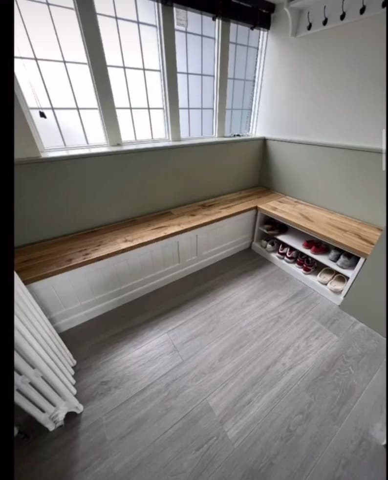 May include: A built-in wooden bench with a white base and a natural wood top. The bench is positioned beneath a large window and includes a shoe storage compartment. The floor is a light gray wood-look laminate.
