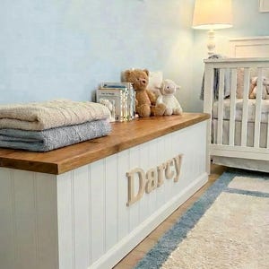 May include: A white and wood-toned storage bench with the name "Darcy" in wooden letters. The bench is in a nursery setting, with a crib, framed animal art, and a rug. Soft blankets and stuffed animals are on top.