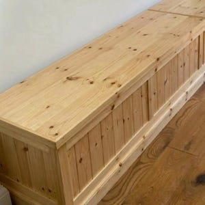 May include: A long, rectangular wooden bench with a light-coloured finish. The bench has a paneled front and a flat top. The wood grain is visible, and the bench appears to be made of pine. The bench is approximately 183 cm long.