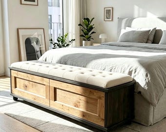 May include: A rectangular storage bench featuring a natural light wood finish with dark wood trim and a cream-colored tufted upholstered seat cushion. The bench is positioned at the foot of a bed and rests on small, dark, rounded feet.