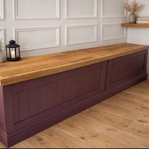 May include: A long, rectangular storage bench with a stained wood top and a deep burgundy base. The bench has a paneled front and sits on a wooden floor. A black lantern with a lit candle sits on top.