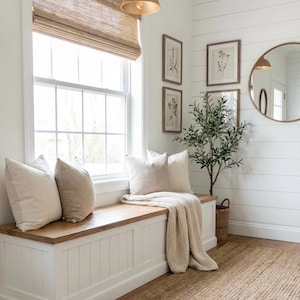 May include: A cozy interior scene featuring a white bench with a wooden top, adorned with beige pillows and a knitted throw. A woven rug and a small potted plant add warmth. The room has a large window with a woven shade and framed botanical prints.