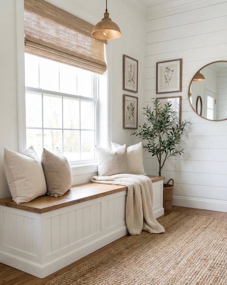 May include: A cozy interior scene featuring a white bench with a wooden top, adorned with beige pillows and a knitted throw. A woven rug and a small potted plant add warmth. The room has a large window with a woven shade and framed botanical prints.