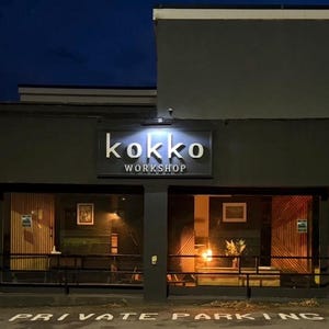 May include: Exterior view of a building at night with the name "kokko WORKSHOP" in illuminated letters above the entrance. The building has large windows and a dark gray facade. "PRIVATE PARKING" is painted on the pavement.