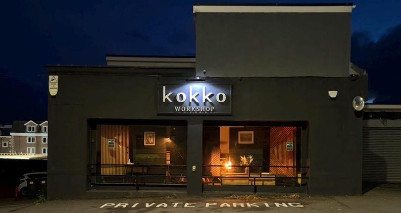 May include: Exterior view of a building at night with the name "kokko WORKSHOP" in illuminated letters above the entrance. The building has large windows and a dark gray facade. "PRIVATE PARKING" is painted on the pavement.