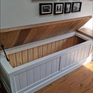 May include: A white and wood-toned storage chest with an open lid. The chest has a rectangular shape with vertical paneling on the front. The lid is made of wood with a diagonal pattern. The interior is light-colored wood.