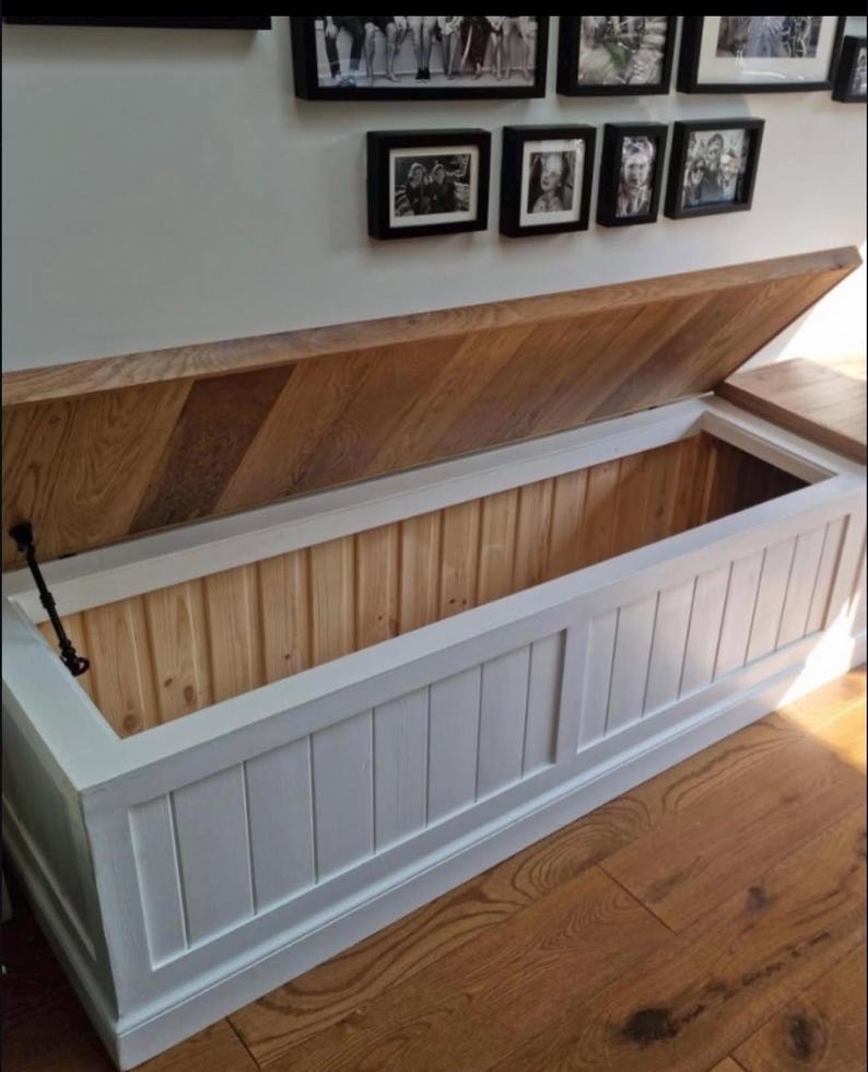 May include: A white and wood-toned storage chest with an open lid. The chest has a rectangular shape with vertical paneling on the front. The lid is made of wood with a diagonal pattern. The interior is light-colored wood.