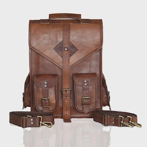 May include: A brown leather backpack featuring a top handle, a central vertical strap with a buckle, and two front pockets with buckled flaps. The bag includes two detachable shoulder straps with metal clips positioned at the base of the item.