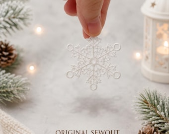 Può includere: Un ornamento a forma di fiocco di neve in pizzo bianco con motivi intricati e delicati, dotato di sei anelli circolari alle estremità dei rami. Il fiocco di neve è tenuto davanti a uno sfondo neutro, evidenziando il ricamo dettagliato.