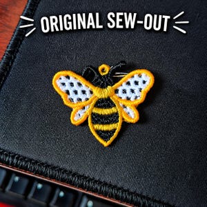 Puede incluir: Parche bordado de abeja con detalles amarillos, negros y blancos. La abeja tiene un contorno amarillo, un cuerpo a rayas negras y amarillas y alas blancas con puntos negros. El texto "ORIGINAL SEW-OUT" está arriba.