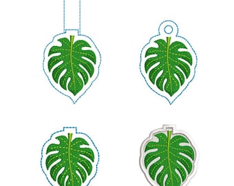 Può includere: Quattro disegni di foglie di monstera ricamate in verde con cuciture delle venature gialle. Tre disegni presentano contorni tratteggiati blu per il taglio, inclusi uno con una linguetta rettangolare e uno con un foro circolare. Un disegno ha un bordo bianco finito.