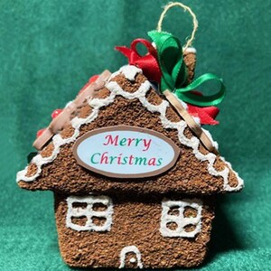 May include: A gingerbread house Christmas ornament with white icing trim, red and green ribbon, and the words "Merry Christmas" on an oval sign. The ornament has two white window designs and a small chimney.
