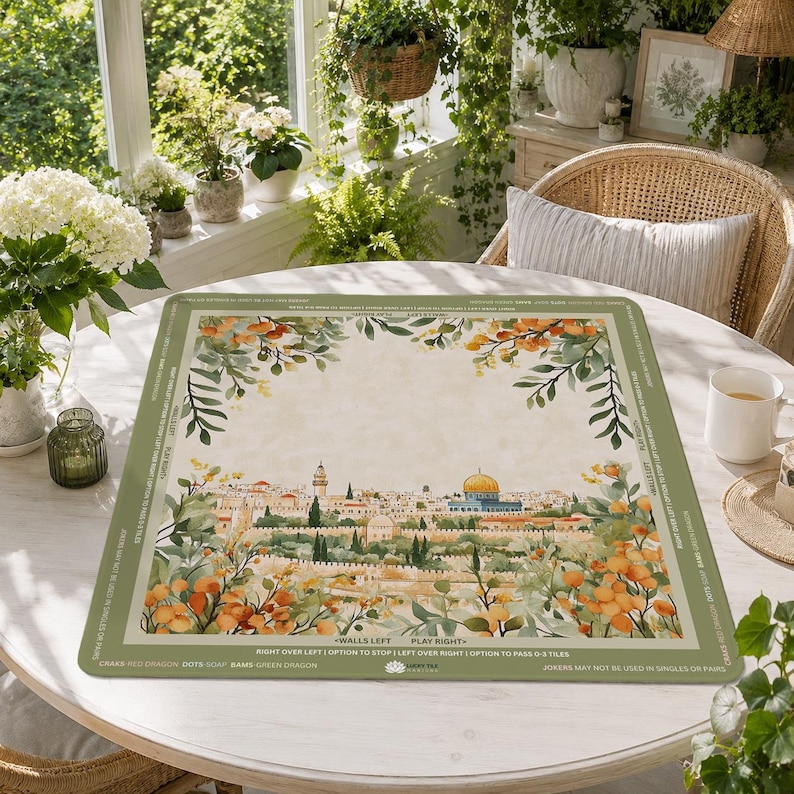 May include: A rectangular game mat featuring a watercolor illustration of a cityscape with a golden dome and surrounding buildings. The mat has a green border with text and floral accents. The mat is on a white table.