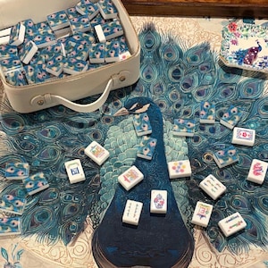 May include: A mahjong set featuring white tiles with blue and green peacock feather patterns, stored in a white rectangular carrying case. Several tiles are scattered on a surface with a large peacock print, alongside a small square dish.