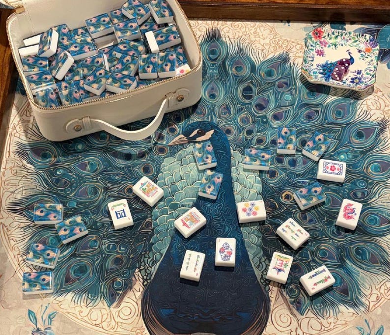 May include: A mahjong set featuring white tiles with blue and green peacock feather patterns, stored in a white rectangular carrying case. Several tiles are scattered on a surface with a large peacock print, alongside a small square dish.
