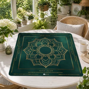 May include: A square, emerald green game mat with a gold mandala design in the center. The mat has gold text around the edges with game instructions. The mat is on a white table in a bright room with plants.
