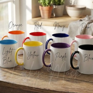 May include: A collection of white ceramic mugs with colored interiors and handles. The mugs feature the colors orange, red, navy, pink, light blue, yellow, purple, and black. Each mug has a handwritten word on the side.