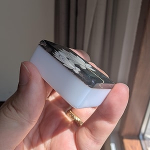 May include: A small rectangular mahjong tile with a smooth white base and a black top surface. The black top features a white engraved graphic of a stylized flower with multiple petals. The tile has rounded edges and a polished finish.
