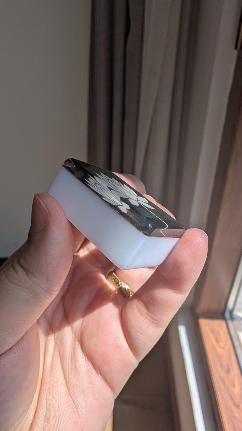 May include: A small rectangular mahjong tile with a smooth white base and a black top surface. The black top features a white engraved graphic of a stylized flower with multiple petals. The tile has rounded edges and a polished finish.