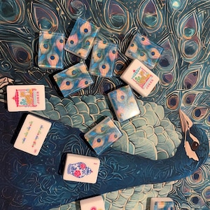 May include: A collection of rectangular mahjong tiles featuring various designs, including peacock feather patterns, floral vases, colorful circles, and two tiles labeled JOKER with a giraffe illustration, scattered across a peacock feather print.