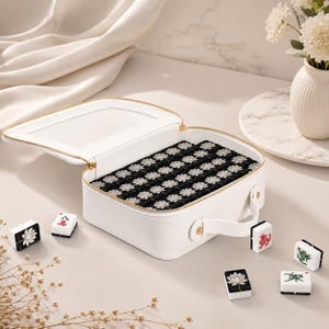 May include: A white rectangular carrying case with a gold zipper and handle, filled with black mahjong tiles featuring white lotus flower designs. Several individual mahjong tiles with lotus, red character, and green dragon symbols are placed nearby.