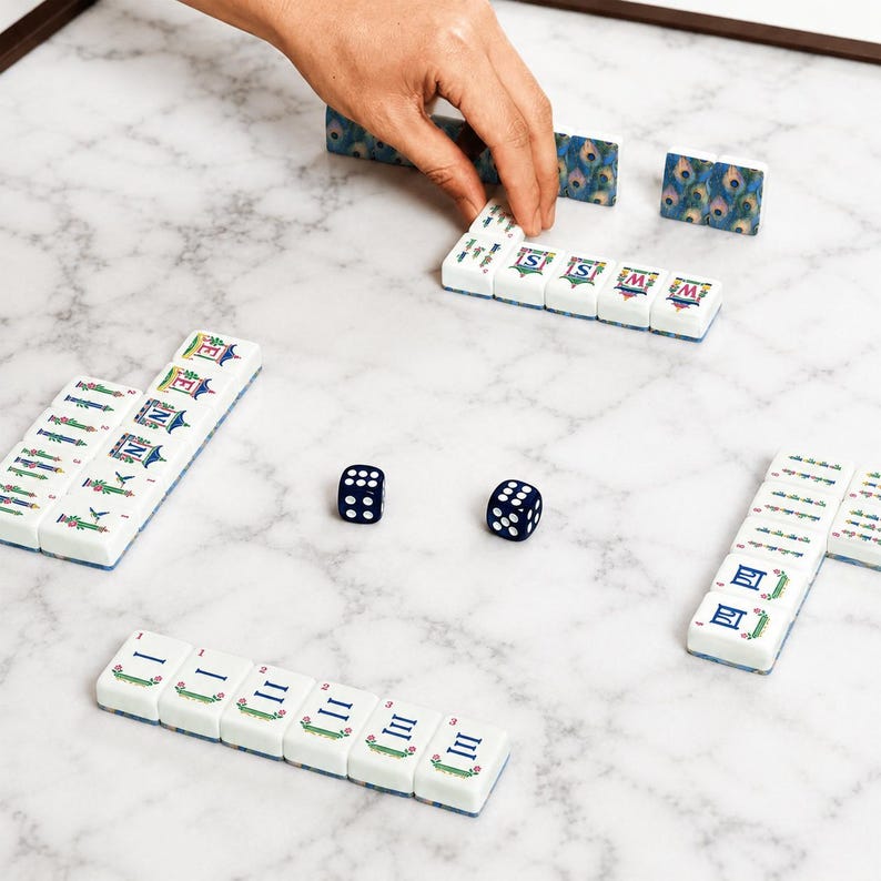 May include: A mahjong set featuring white tiles with colorful illustrations of pagodas, peacock feathers, and Chinese numerals. The set includes two dark blue dice with white pips, arranged on a white surface with grey marble veining patterns.