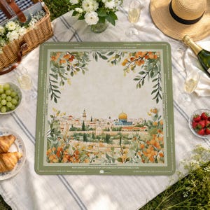 May include: A square, green-bordered game board featuring a watercolor illustration of a city with a golden dome, surrounded by floral elements. The board is set on a picnic blanket with food and drinks.
