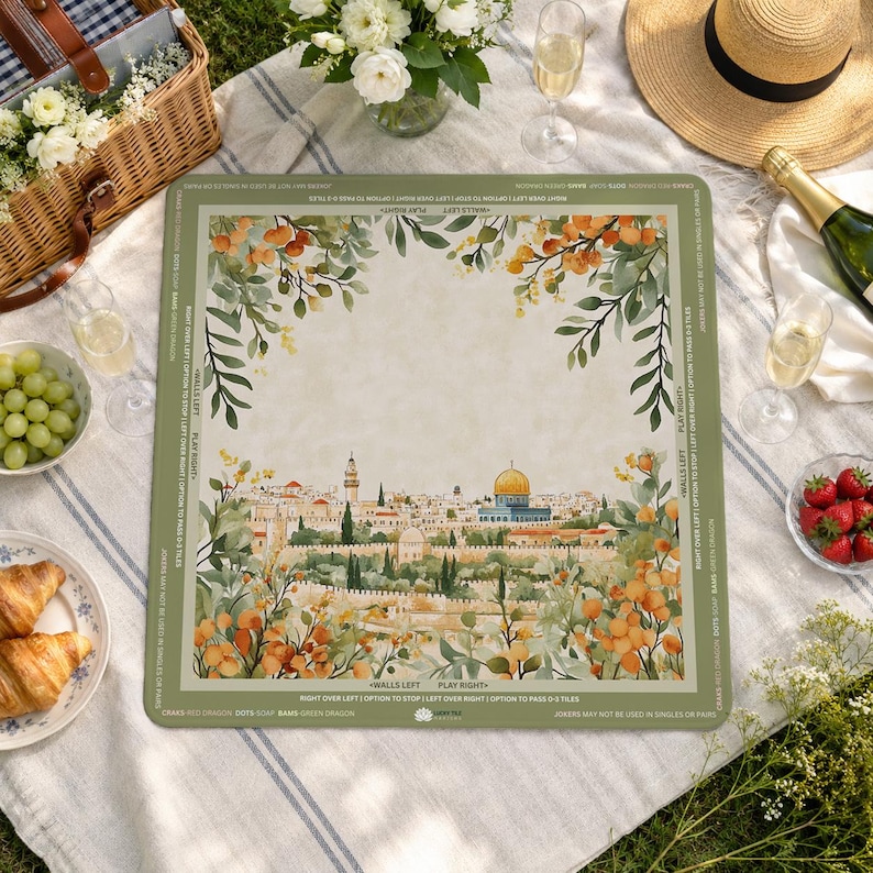 May include: A square, green-bordered game board featuring a watercolor illustration of a city with a golden dome, surrounded by floral elements. The board is set on a picnic blanket with food and drinks.