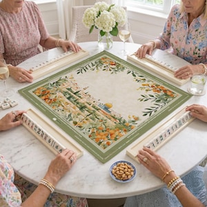 May include: A round table with four people playing a tile-based game. The game board is a square mat with a green border and a watercolor illustration of a building and orange fruit. Each player has a rack of tiles.