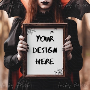 May include: A framed rectangular sign with the text "YOUR DESIGN HERE" in bold black letters. The frame is dark brown, and the sign has spiderweb and spider decorations. The person holding the sign is wearing a black outfit and bat wings.