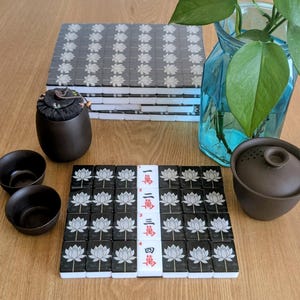 May include: A set of black and white mahjong tiles featuring lotus flower designs, arranged in a grid. The center column displays tiles with red Chinese characters. Included are two dark brown ceramic tea cups, a lidded tea pot, and a tea canister.