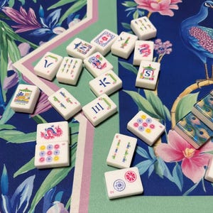 May include: A collection of white rectangular mahjong tiles featuring colorful engraved symbols, including dragons, flowers, numbers, and letters. The tiles are scattered across a blue and green surface decorated with a peacock and floral pattern.
