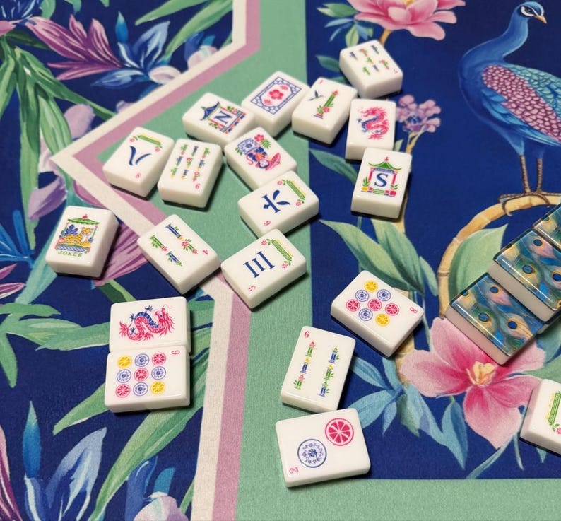 May include: A collection of white rectangular mahjong tiles featuring colorful engraved symbols, including dragons, flowers, numbers, and letters. The tiles are scattered across a blue and green surface decorated with a peacock and floral pattern.