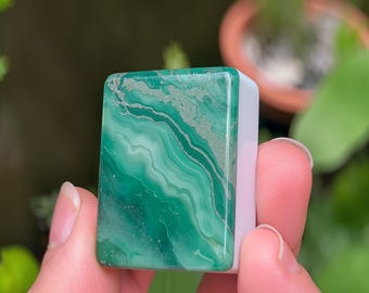 May include: A rectangular mahjong tile featuring a smooth, glossy green surface with intricate, swirling white and dark green marbled patterns. The tile has a solid white base and rounded corners, displaying a polished finish on its front face.