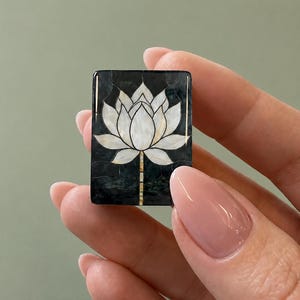 May include: A rectangular inlay piece featuring a white mother-of-pearl lotus flower design set against a dark, mottled black background. The lotus petals are segmented with thin dark lines, and the stem extends downward in a vertical pattern.