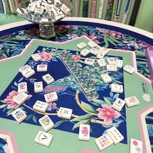 May include: A Mahjong set featuring white tiles with colorful illustrations and characters, scattered across a table mat with a blue and green peacock and floral pattern. A glass bowl filled with additional tiles sits on the table.