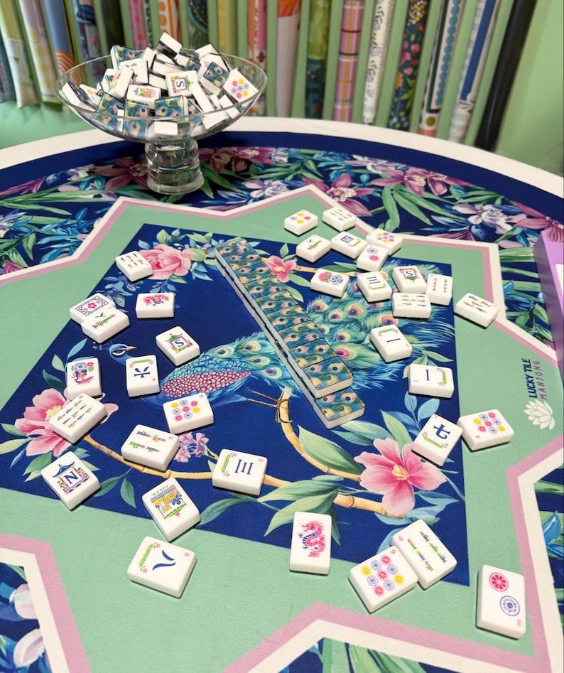 May include: A Mahjong set featuring white tiles with colorful illustrations and characters, scattered across a table mat with a blue and green peacock and floral pattern. A glass bowl filled with additional tiles sits on the table.