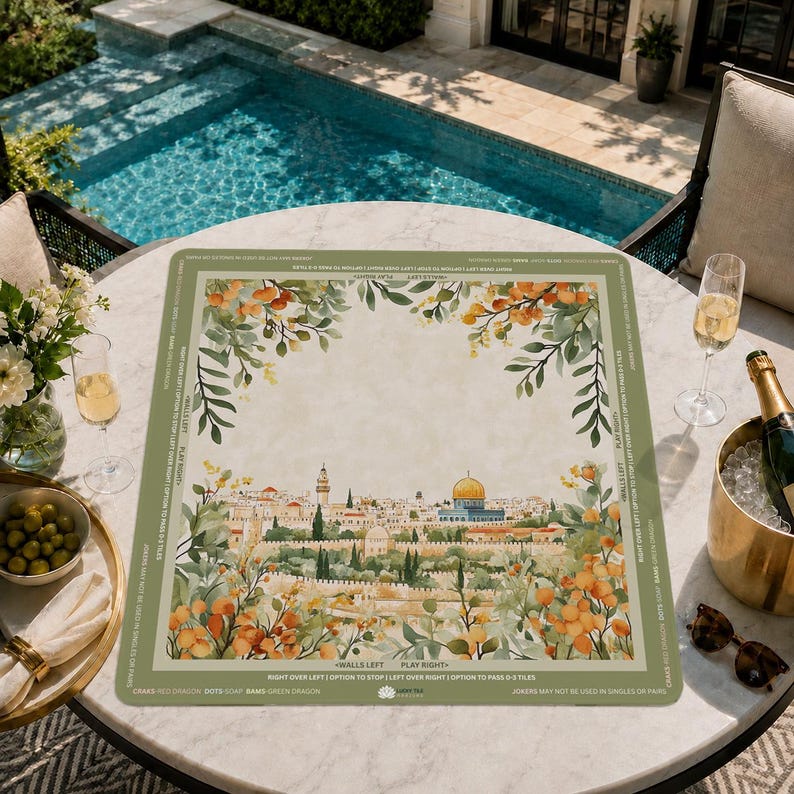 May include: A rectangular placemat with a watercolor illustration of a cityscape, featuring a golden-domed building and surrounding buildings. The placemat has a green border with text and floral accents. The placemat is on a round table with drinks and snacks.