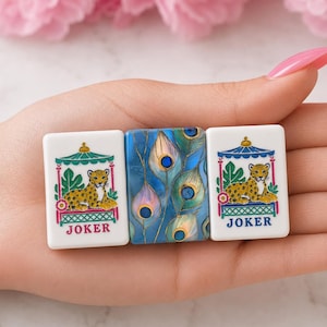 May include: Three rectangular mahjong tiles. The two outer tiles are white with a graphic of a leopard resting under a teal and pink pavilion, with the word JOKER printed in red or blue. The center tile features a blue peacock feather pattern.