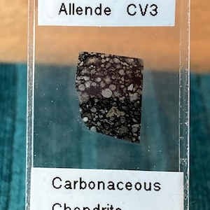 May include: A small, irregularly shaped piece of the Allende CV3 carbonaceous chondrite meteorite, displayed in a clear plastic holder. The meteorite has a dark, speckled appearance with lighter-colored inclusions. Text on the holder reads "Allende CV3" and "Carbonaceous Chondrite."