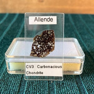 Peut inclure: Un échantillon de la météorite d'Allende, une chondrite carbonée CV3, présenté dans un boîtier en plastique transparent. Le fragment de météorite est brun foncé avec des inclusions blanches. L'étiquette indique "Allende" et "CV3 Carbonaceous Chondrite."