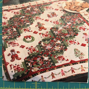 May include: A quilted fabric featuring a Christmas theme with gingerbread people, wreaths, and trees in red, green, and white. The quilt is positioned above a yellow measuring tape showing a width of approximately 7.5 inches across the bottom edge.
