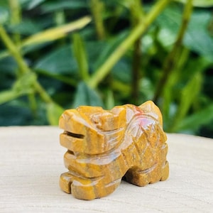May include: A small, hand-carved tiger's eye gemstone figurine shaped like a mythical creature. The stone features a golden-brown colour with natural chatoyant bands and a polished, smooth surface, depicting a stylised animal with defined facial features.