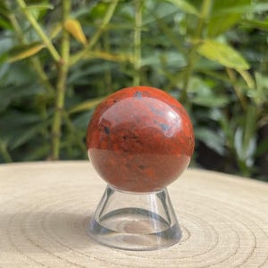 May include: A polished, spherical red jasper stone with dark brown and black inclusions, resting on a clear, conical acrylic display stand. The stone features a smooth, glossy surface with natural colour variations throughout its round form.